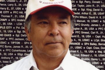 Dugan Aguilar stands in front of a black slab etched with white names of deceased veterans