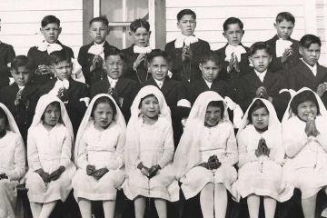 A black and white photograph of Indigenous schoolchildren with hands clasped as if in prayer. Girls dressed in white sit in the front row, while boys dressed in all black stand behind them.