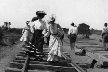 A black and white photo of two women walking on wooden train tracks. 