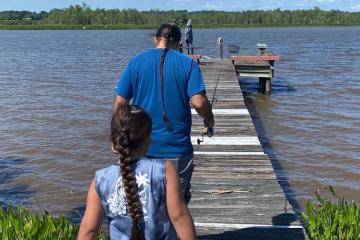 A father and son holding fishing poles walk down a dock toward a river