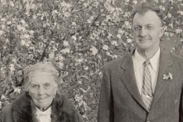 Isabel Meadows in a coat with a fur collar stands next to John Peabody Harrington wearing a suit in front of a tree