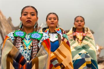 Three jingle dress dancers pose on a fallen tree log on the foggy shore.