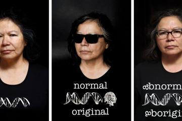 Three photographs of artist Shelley Niro wearing a black t-shirt. In the first, her t-shirt displays a strand of DNA. In the second, there is DNA and a figure wearing a headdress, with the words "Normal Original." In the third, the words are changed to "Abnormally Aboriginal."