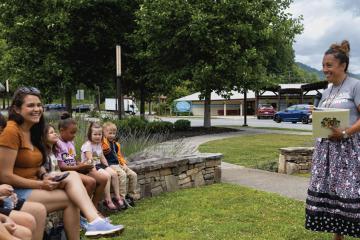 A smiling woman holds a book, facing a semi-circle of children seated on a rock wall outdoors