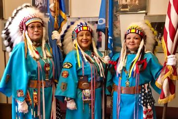Three Native women veterans wearing blue traditional dresses with military decorations and and feather headdresses