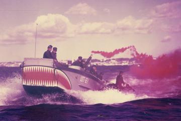 A 1950s photograph of a grey boat with teeth painted on one end carries 8 crew members on board. One crew member is holding a flare releasing red smoke.