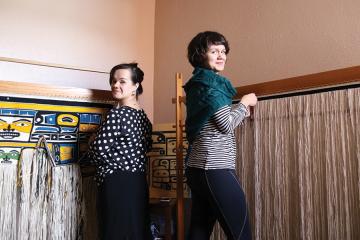 Two women stand back to back, each in front of a weaving loom