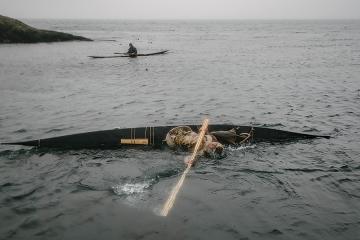 A person extends a paddle while seated in a kayak that has tipped over, in a body of water