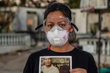 A woman wearing safety goggles and a respirator mask stands in front of the remains of her home holding a photograph