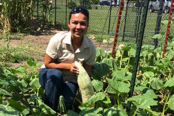 A Cherokee woman crouches in a garden, holding a squash