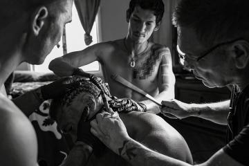 A tattoo artist applies a tattoo while two assistants stretch the skin of the recipient