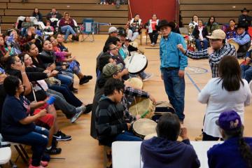 Crowd of people seated in a gymnasium observe participants playing a handgame. 