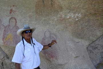 Person in uniform and white western hat points at paintings of people on a rock wall.