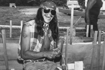 Person in aviator sunglasses, flannel vest, and bandana splits roots for a canoe-building workshop.