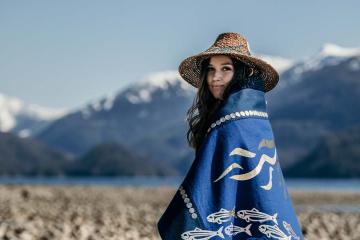 An Indigenous woman stands along a beach in a blue cloak depicting herrings.