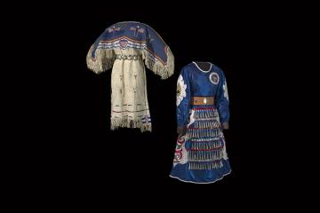 Two dresses stand back to back on mannequins: the dress on the left depicts American flags and stripes made with tanned hide. The dress on the right is constructed with blue fabric and ordained with metallic cones, military patches, and sequins. 
