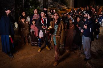 Actors on stage depict the Trail of Tears: a large group of Cherokee peoples are forcefully led by two soldiers.
